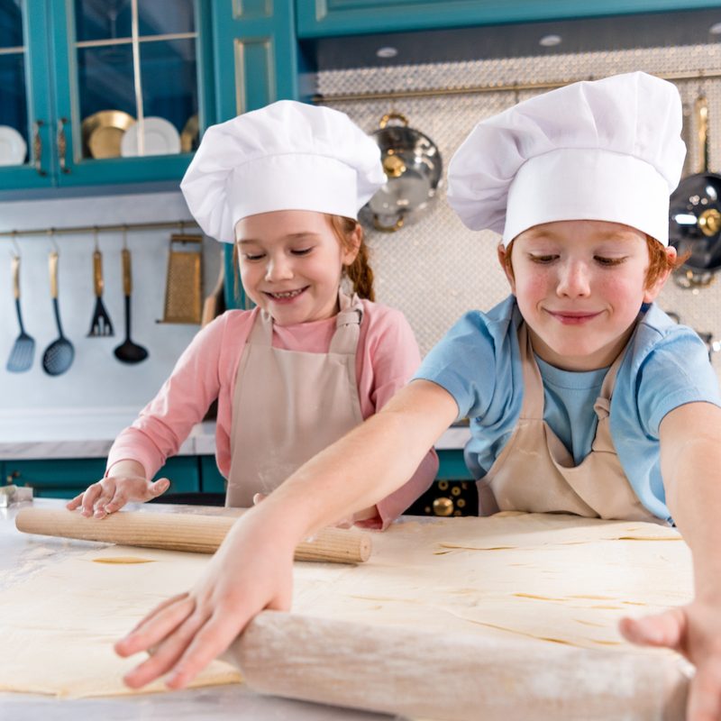 happy little kids in chef hats and aprons rolling dough in kitchen